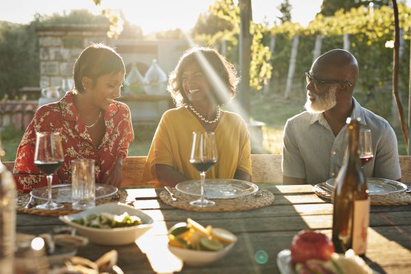 Three people sitting at an outdoor table, smiling and talking, with wine glasses and plates in front of them during a sunny day.