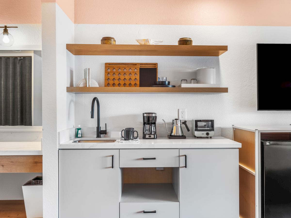 A modern kitchenette with open shelves, a sink, coffee maker, and other appliances. There's a flat-screen TV next to a mini-fridge.