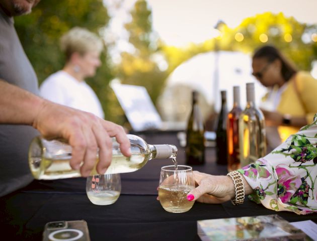 A person is pouring white wine into a glass held by someone with a floral shirt at an outdoor gathering with bottles of wine on the table.