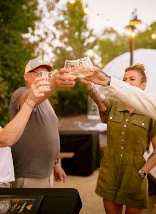 A group of people are smiling and raising their glasses in a toast, enjoying an outdoor gathering with greenery and lights in the background.