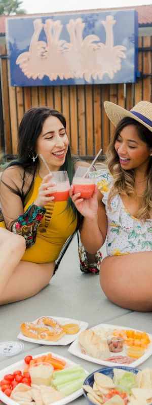 Two women are sitting by a pool, enjoying drinks and snacks with plates of food spread out in front of them, on a sunny day.