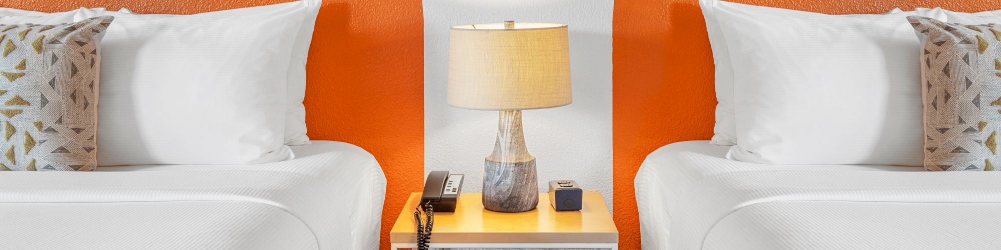 Two beds with white linens and decorative pillows are separated by a small nightstand holding a lamp, phone, and other small items.