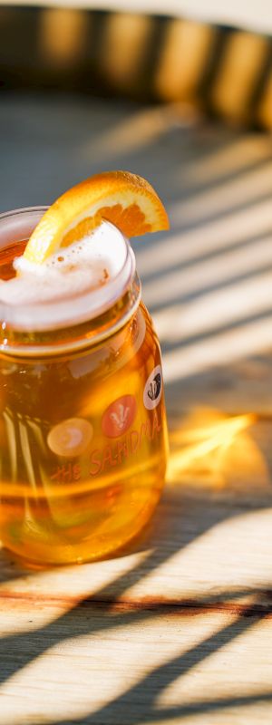 The image shows a glass of beer with a slice of orange garnish placed on a wooden surface with shadows cast by striped lighting.