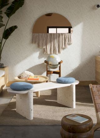 A cozy, modern living room with wooden furniture, a plant, books, globe, round mirror, wicker chair, and eclectic decor against a light-colored wall.