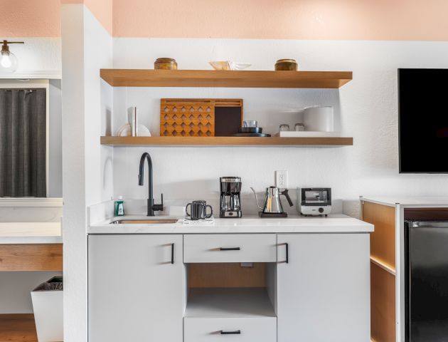 A kitchenette with a sink, coffee maker, toaster, and open shelves stocked with containers, a microwave, and a refrigerator.