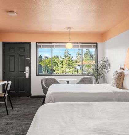 A hotel room with two beds, a desk, chairs, and a large window looking outside. The decor includes an orange circular accent above the beds.