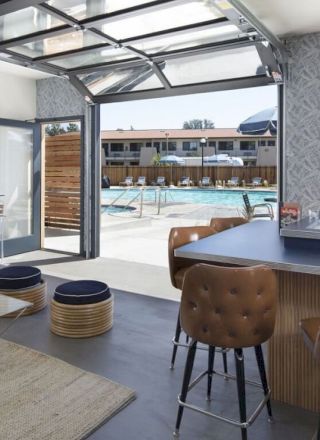 The image shows a modern interior with a bar area, seating, and a large open window overlooking a pool with lounge chairs outside.