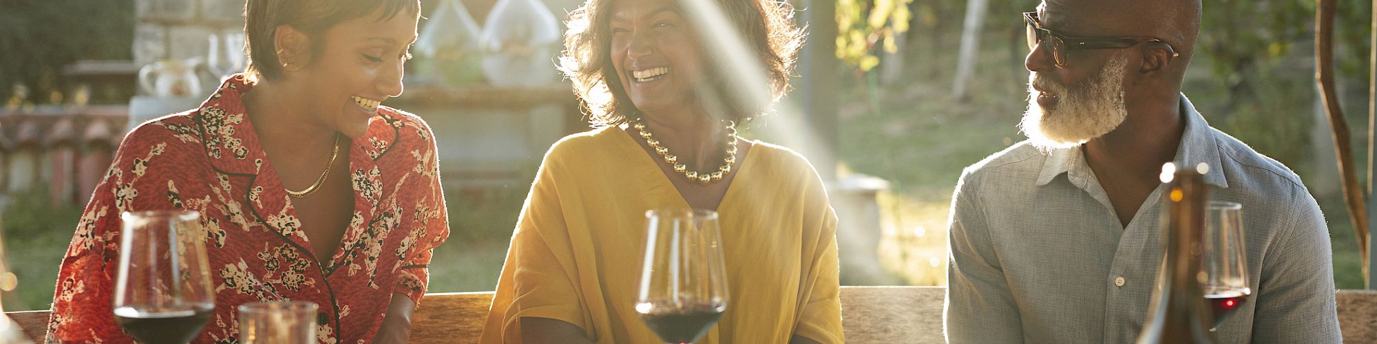 Three people are sitting outdoors at a table, enjoying a meal with wine and looking happy as they share a conversation in the sunlit setting.