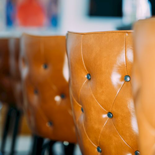 The image shows a row of brown leather chairs with button tufting in a blurred background setting, possibly in a dining area.