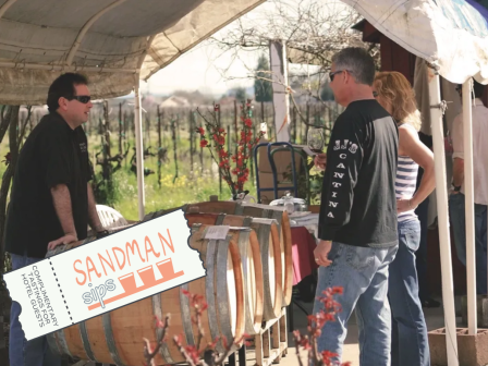 Three people are talking at an outdoor market stand with barrels, blooming tree branches, and a 