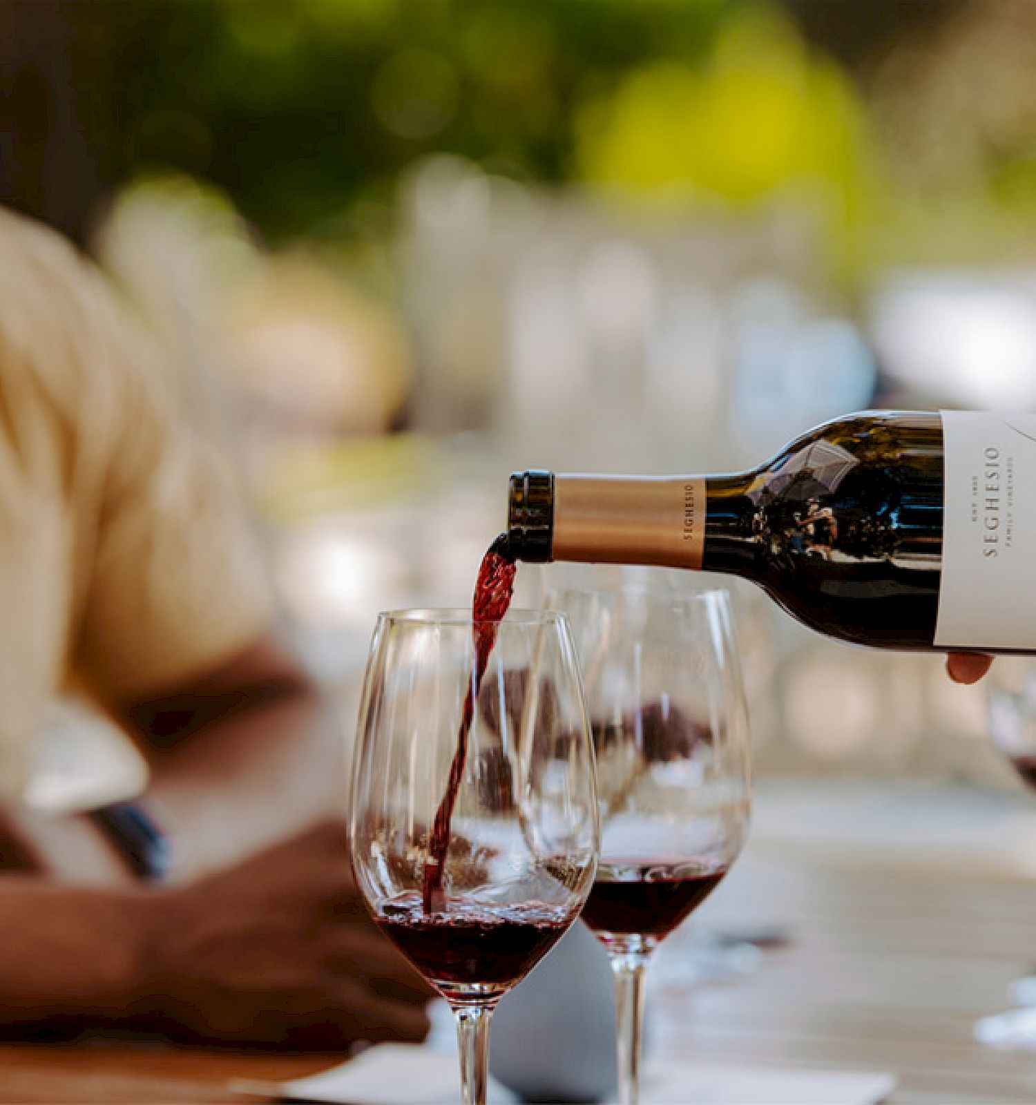 Someone is pouring red wine from a bottle into several glasses on a table, with people in the background sitting outdoors.